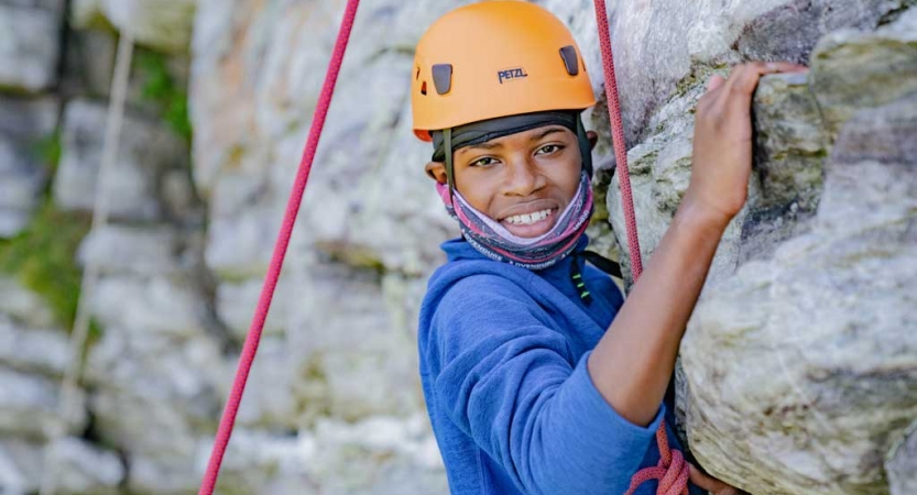 a young person wearing a helmet and secured by ropes pauses rock climbing to smile at the camera